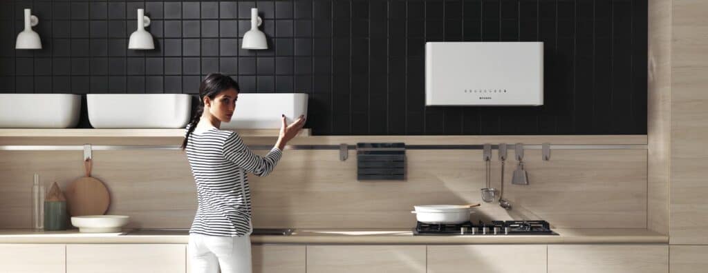 A woman in a modern kitchen, standing near a counter with cooking utensils, a stove, and white storage units against a black tiled wall.