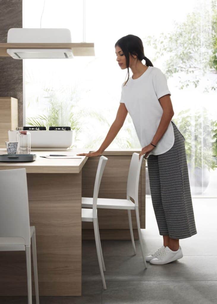 A woman in a white t-shirt and patterned pants is standing in a modern kitchen, leaning on the counter.