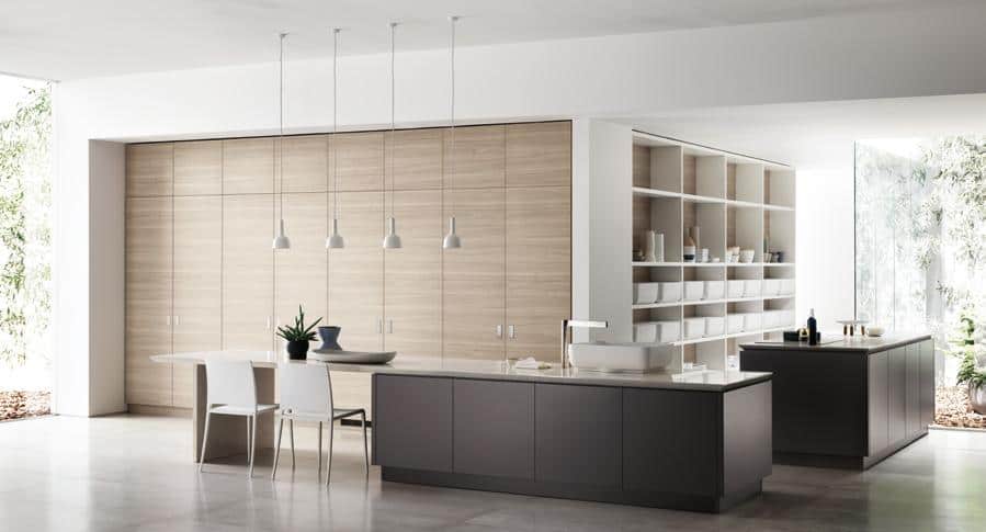 Modern kitchen with light wood cabinetry, pendant lighting, and two islands; one with a sink and the other with shelves and decorative items. Large windows provide natural light.