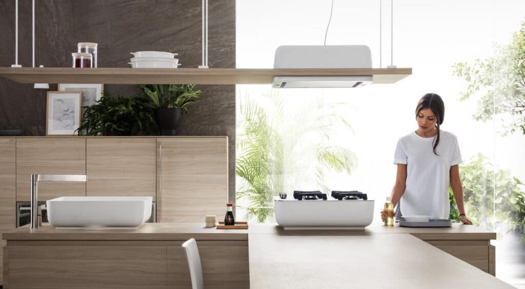 Modern kitchen with wooden cabinetry, a countertop sink, plants, and a person standing by a stove.