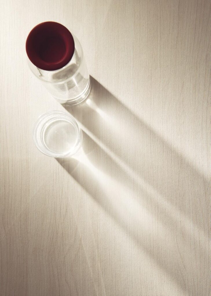 Glass bottle with a red lid and a glass cup casting long shadows on a light wooden surface.