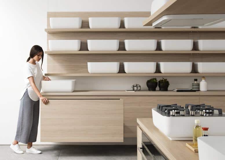 A woman stands in a modern kitchen with wooden cabinets and shelves, featuring white rectangular containers and a stovetop with burners.