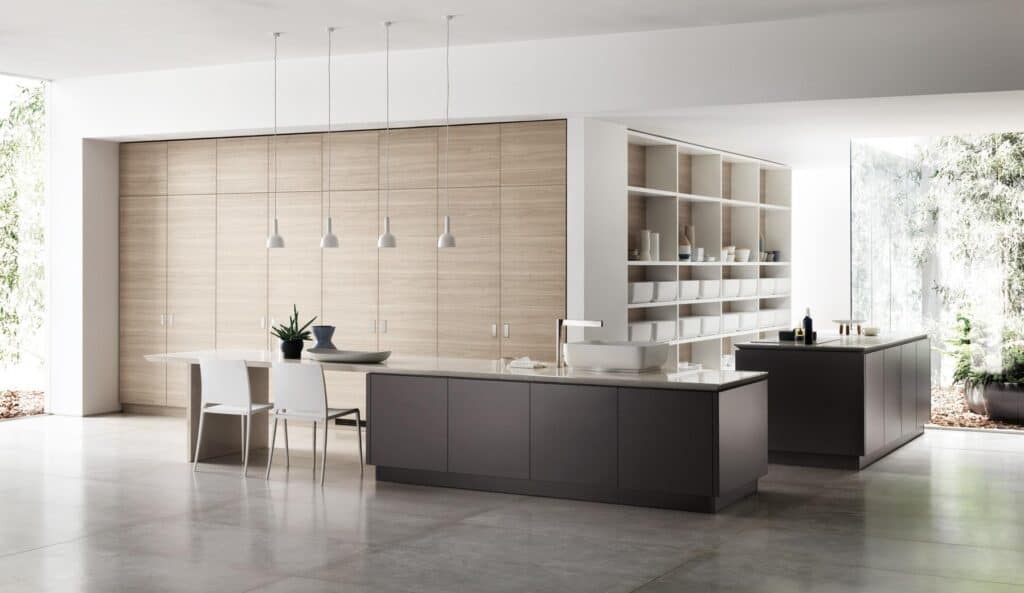 Modern kitchen interior with minimalist design, featuring a large island, white chairs, pendant lights, light wood cabinetry, open shelving, and large windows showcasing greenery outside.