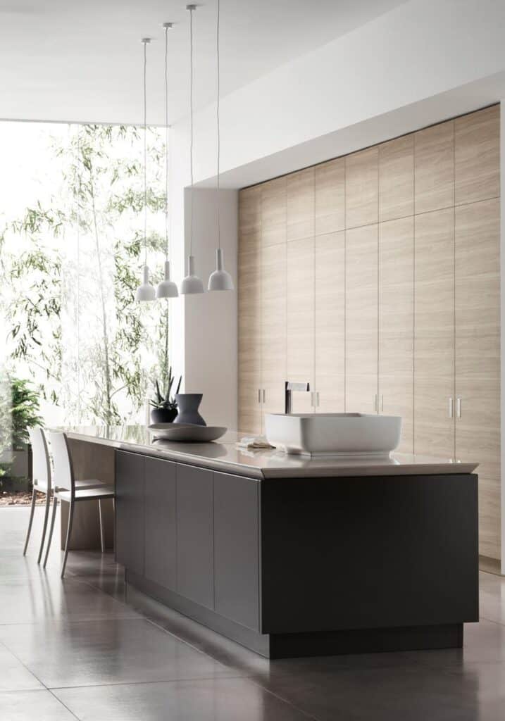 A modern kitchen interior with a large island, dark cabinetry, a countertop sink, and hanging pendant lights, set against light wood cabinets and large windows.