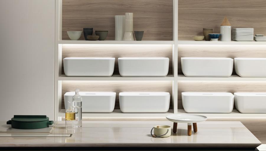A modern kitchen with wooden shelves displaying white rectangular containers, various bowls, and dishware. A countertop features a teapot, a small table, and a bottle.