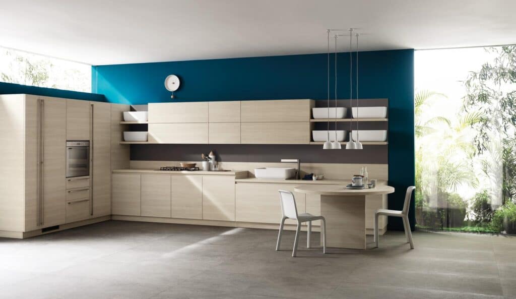 Modern kitchen with light wood cabinetry, teal accent wall, built-in oven, minimalist dining area, pendant lights, and large windows with garden view.