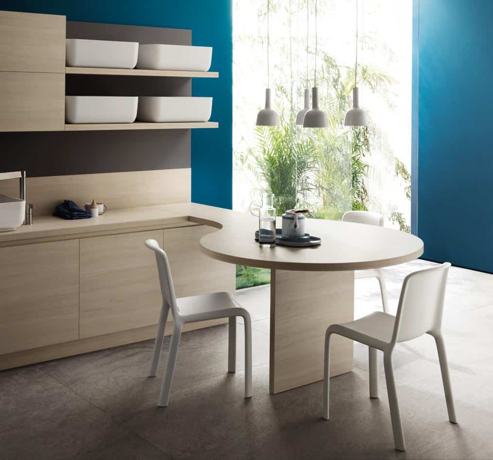 Modern kitchen interior with wooden cabinetry, round dining table, white chairs, pendant lights, and large windows overlooking greenery.