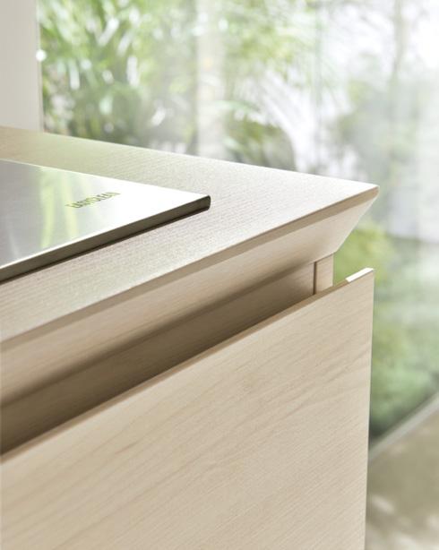 Close-up of a light wooden cabinet with a sleek metal handle and a reflective surface on top, set against a blurred green background.