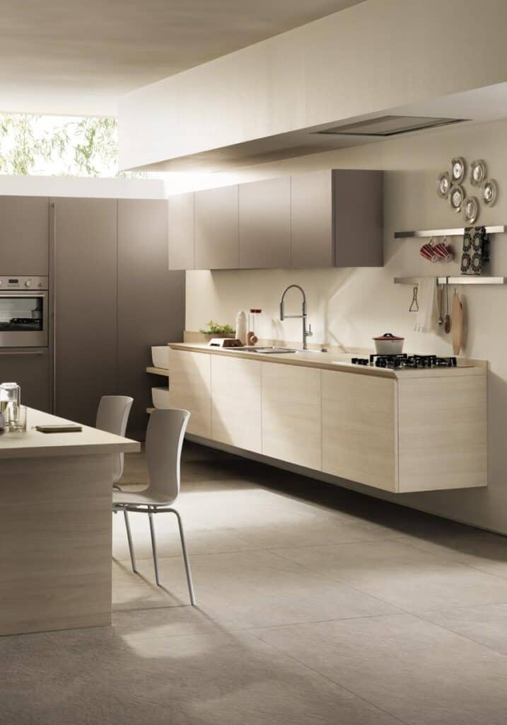 Modern kitchen with light wood cabinets, a built-in oven, a sink, and gas stove. There is a kitchen island with two chairs, shelves with decor, and neutral stone flooring.