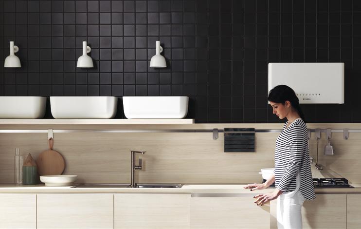 A person in a striped shirt stands in a modern kitchen with a black tiled wall, white cabinets, and a stainless steel faucet.
