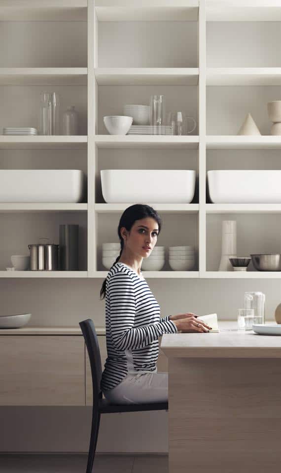 A woman sitting at a table in a modern kitchen with open shelving displaying white dishes and glassware.