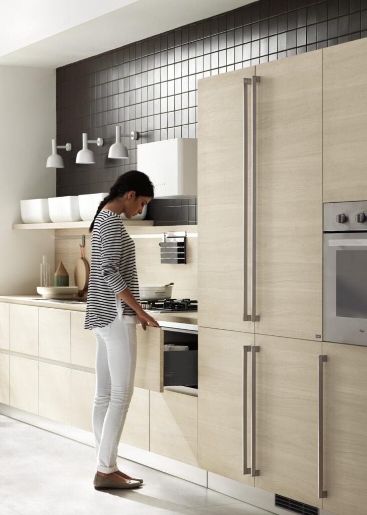 A person is in a modern kitchen with light wood cabinets, opening a drawer near the stove.