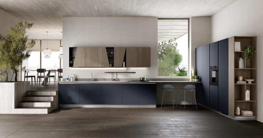 Modern kitchen interior with dark blue cabinets, wooden accents, a sleek countertop, and integrated appliances, adjacent to a dining area with pendant lights and large windows.