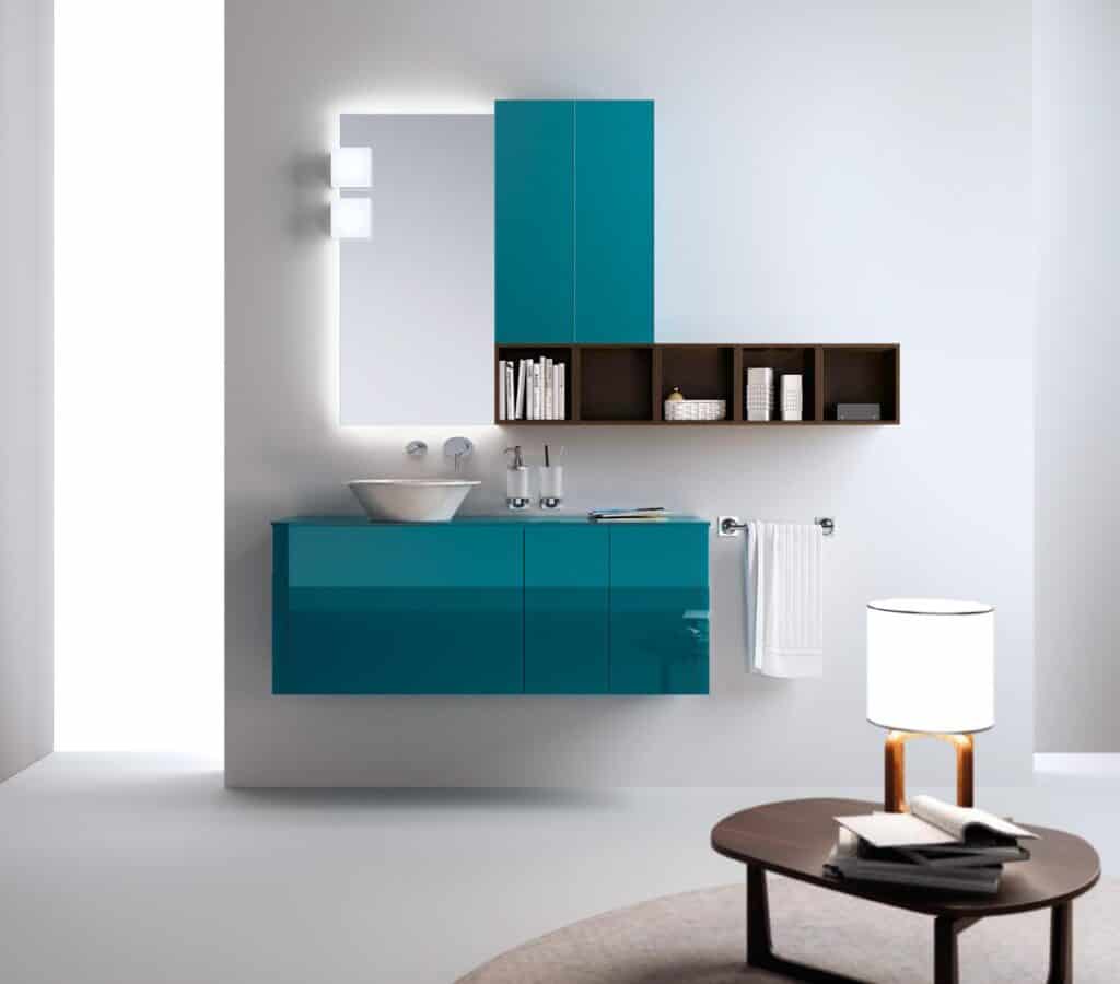 Modern bathroom interior with teal cabinets, a white vessel sink, a backlit mirror, and wall-mounted shelving. A towel hangs on a nearby rack, and there&#039;s a small round table with a lamp.