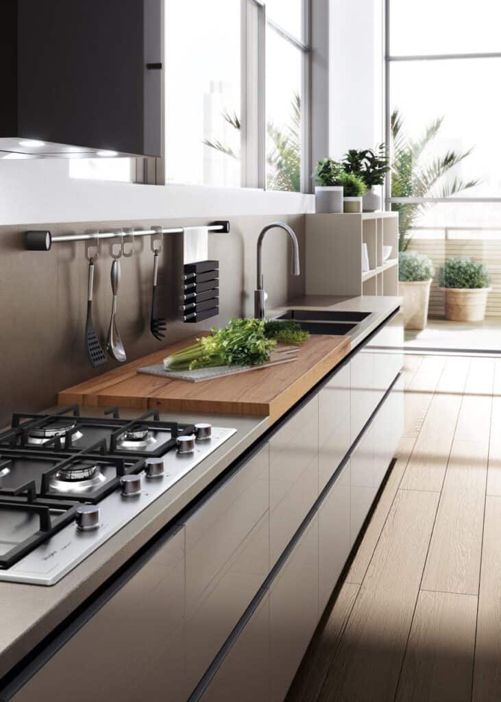 Modern kitchen interior with a gas stove, wooden countertop, sink, hanging utensils, and potted plants by a large window.