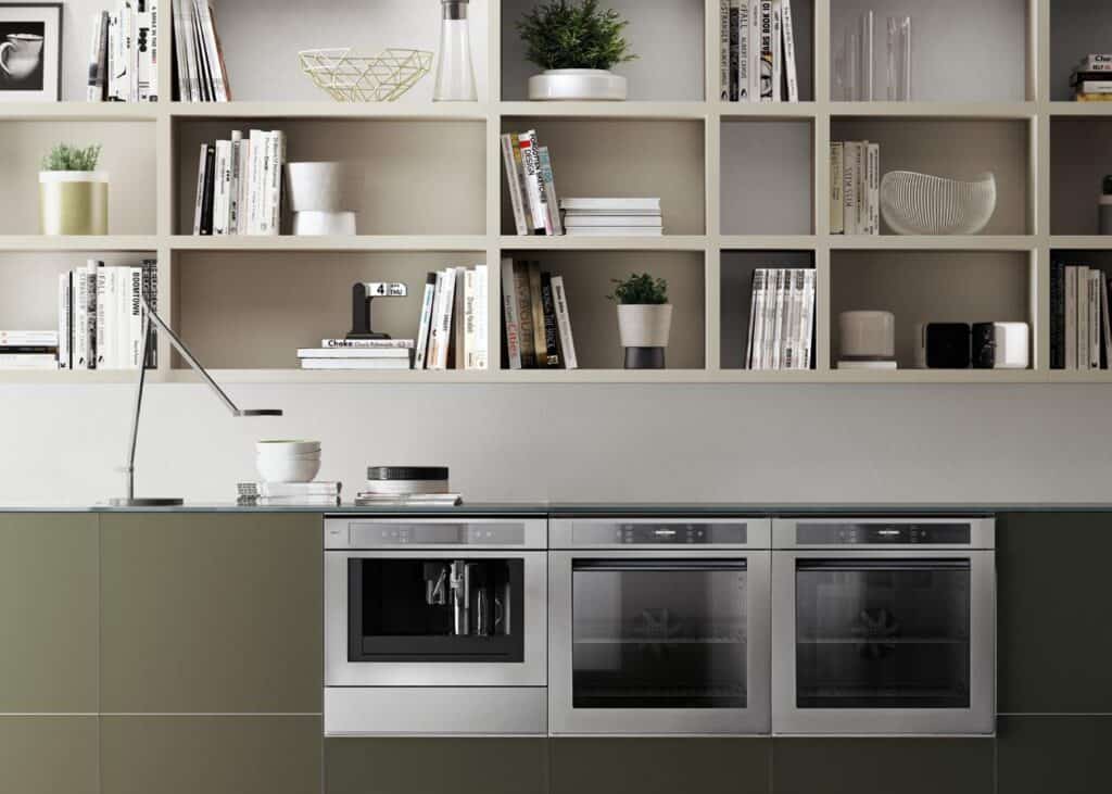 Modern kitchen with built-in appliances, open shelving with books and plants, and a sleek counter with a lamp and bowls.