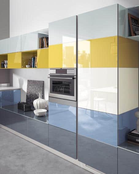 Modern kitchen cabinets with glass doors in blue, yellow, and gray hues, featuring built-in shelves and an oven.