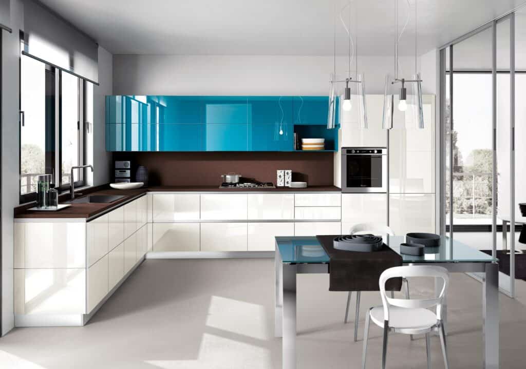Modern kitchen with glossy blue and white cabinets, brown countertops, a glass dining table with white chairs, and pendant lights above.