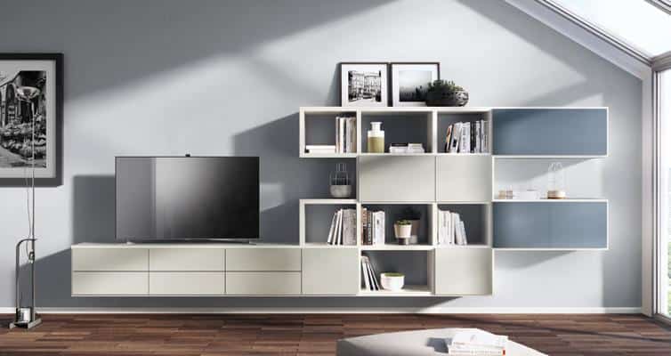 Modern living room with a sleek wall-mounted storage unit, featuring open shelves, drawers, and a flat-screen TV. The floor is wooden, and there&#039;s ample natural light from a slanted ceiling window.