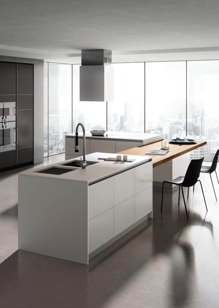 Modern kitchen with minimalist white cabinetry, island with sink, and wooden dining table extension, overlooking a cityscape through floor-to-ceiling windows.