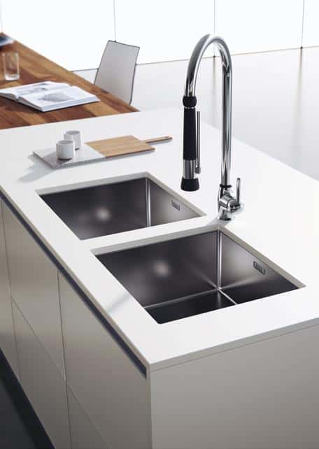 Modern kitchen island with a white countertop, featuring two stainless steel sinks and a tall, sleek faucet. In the background, there&#039;s a wooden table with a chair and a cutting board with cups.