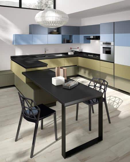 Modern kitchen with black countertops, light blue and olive green cabinets, a sleek black dining table, and two black chairs. A large, white pendant light hangs above the table on light wood flooring.