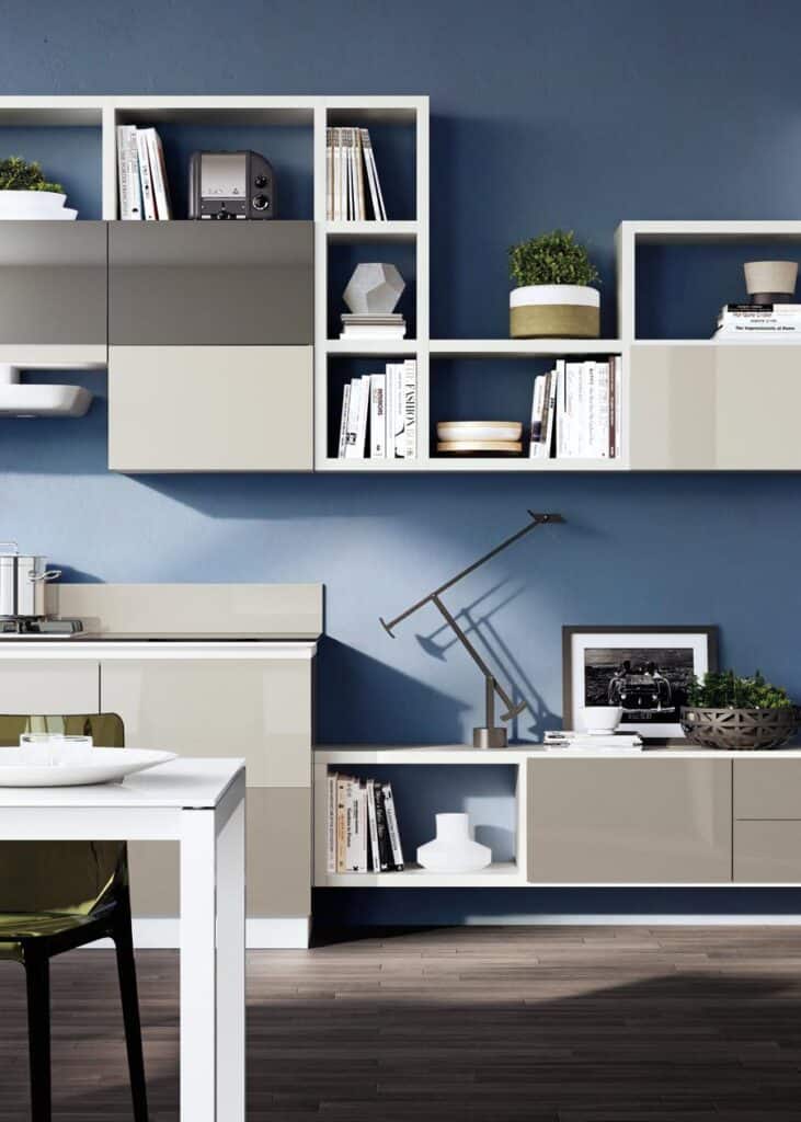 Modern kitchen interior with blue walls, beige cabinets, open shelving with books, potted plants, a photo frame, and a decorative lamp.