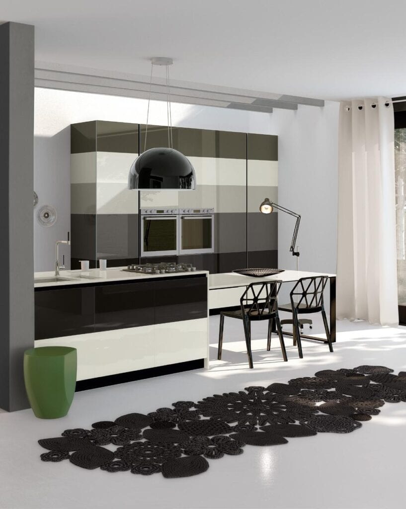 Modern kitchen with sleek black and white cabinetry, a pendant light, a green stool, geometric chairs, and a decorative black rug on a white floor.