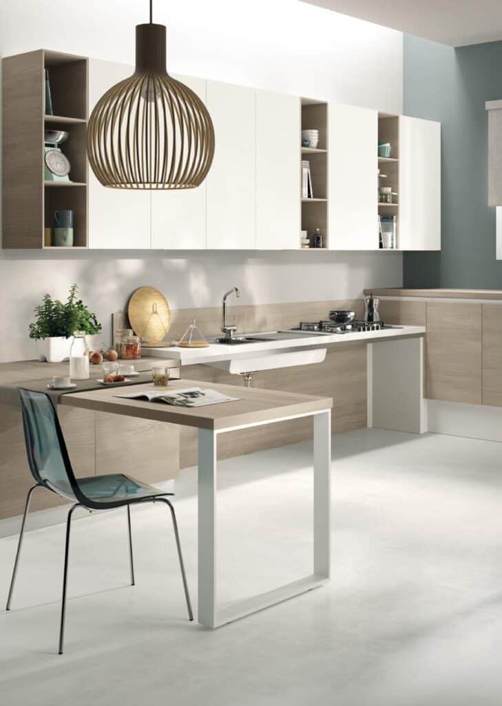 Modern kitchen with light wood cabinetry, a sleek countertop with a built-in sink and stove, and a small dining table with a transparent chair; a stylish pendant light hangs above.