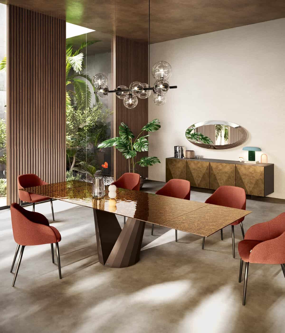 Modern dining room with a long, glossy wooden table surrounded by red upholstered chairs, a spherical chandelier, greenery visible through a window, and a sideboard with a decorative mirror above it.