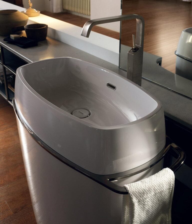 A modern bathroom sink with an oval basin and a sleek, tall faucet. The sink has a towel hanging on a metallic bar, and a mirror is visible in the background.
