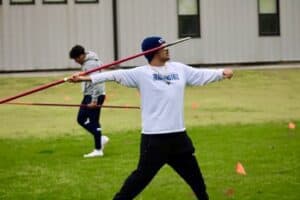 A collegiate athlete practicing javelin throw on a grassy field, wearing a white long-sleeve shirt and a blue cap, with another person practicing in the background.