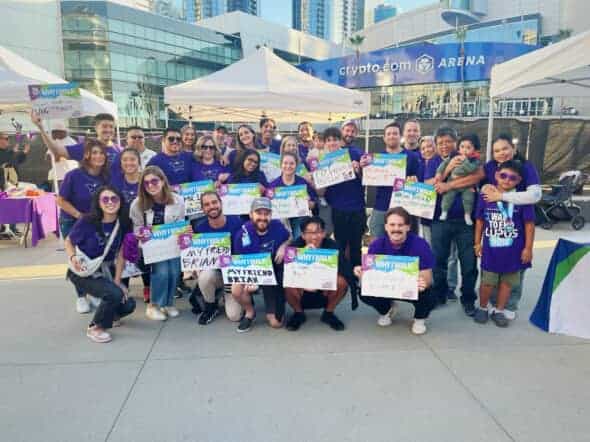 Group photo of individuals wearing purple shirts, gathered outdoors at an event promoting lupus awareness, holding signs and smiling together.
