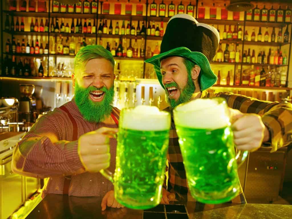 Vibrant St. Patrick's Day celebration with two men wearing green costumes and hats, raising green beers in a lively Irish pub.