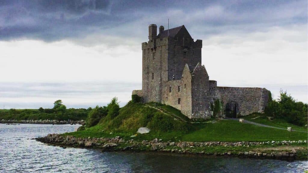 Medieval castle on a lush green hill near water under cloudy sky.
