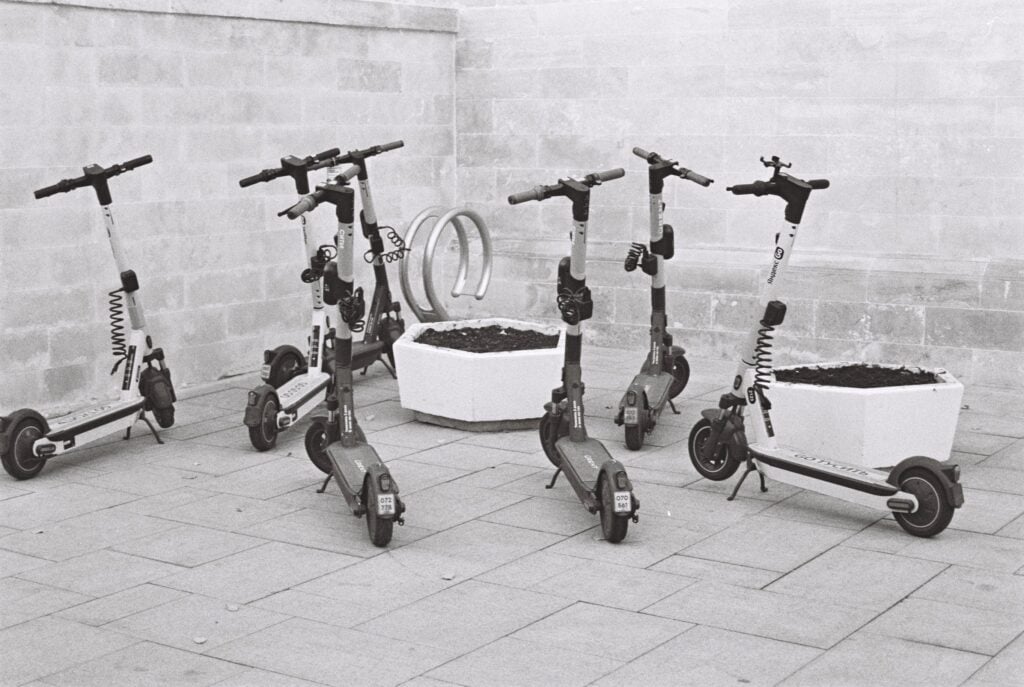 Black and white photo of several electric scooters parked beside concrete planters.