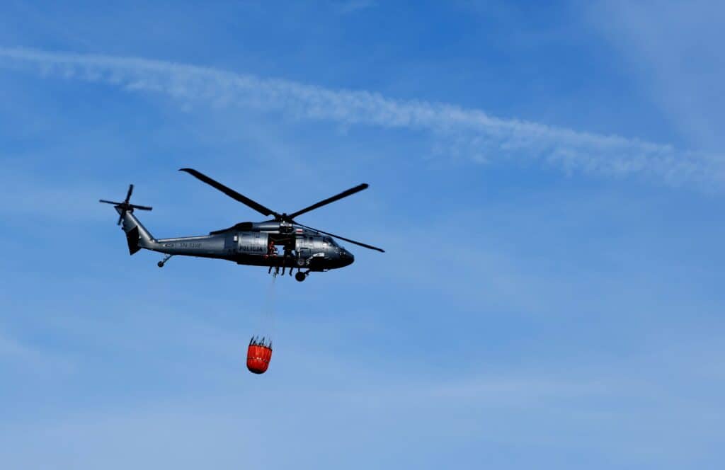 A helicopter flying in the sky with a firefighting bucket, against a clear blue sky.
