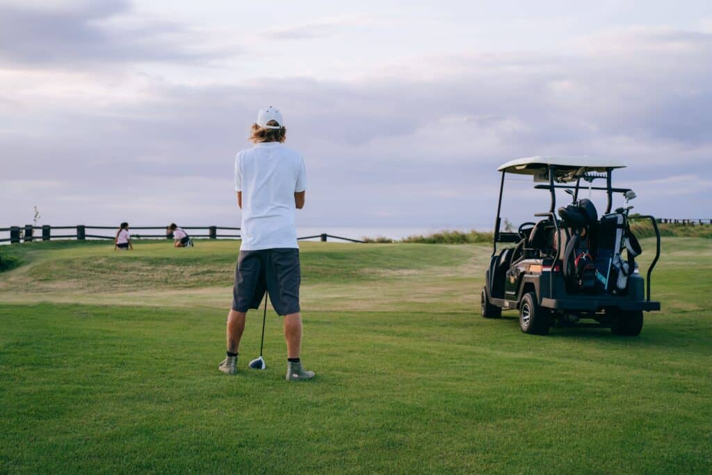 A golfer stands ready to play on a beautiful course with golf cart and clubs nearby.