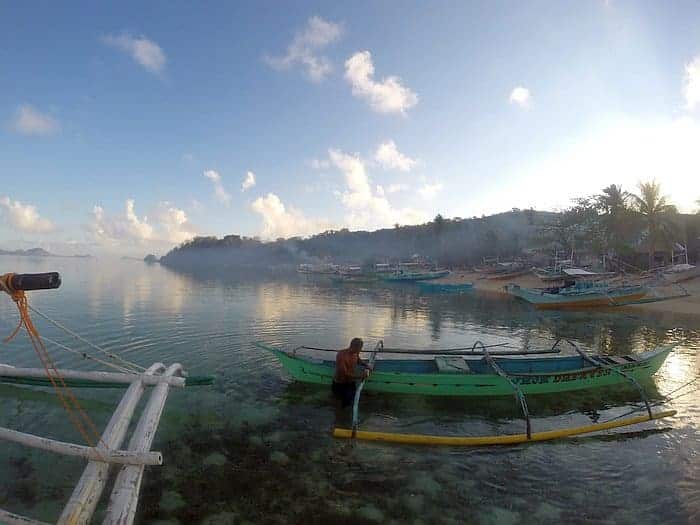 At dawn in El Nido, a man stands serenely on a green boat in shallow, clear water. Several boats, ready for the day's trip, are docked along the shore with palm trees and a hazy sky providing a picturesque backdrop.