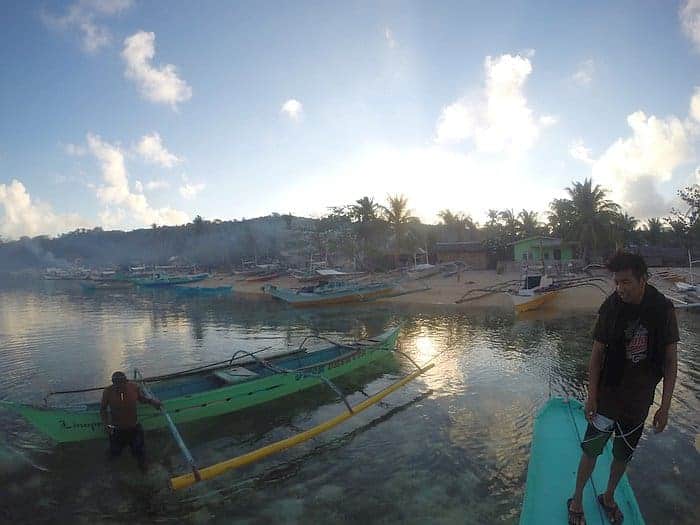 A scenic view of a beach with small boats on the water and the shore, palm trees, and a few people enjoying island hopping as part of a Barangonan boat tour under a bright sky.
