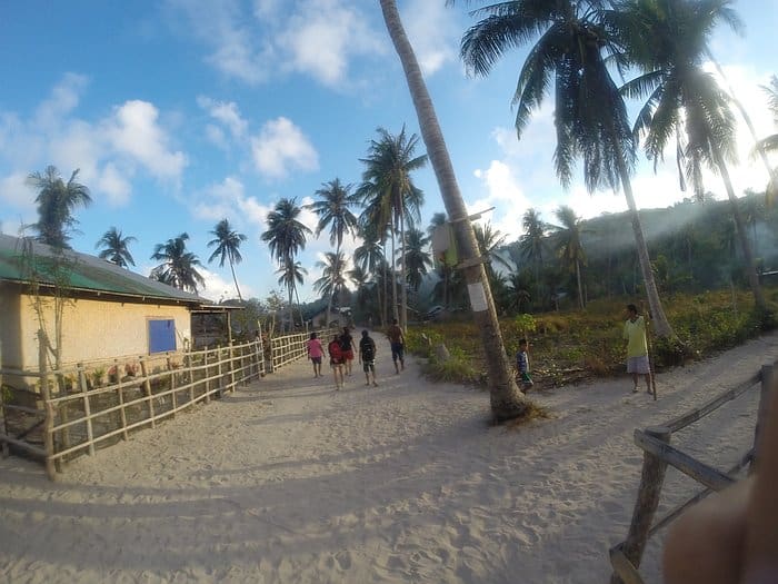 People strolling on a sandy path lined with palm trees and a bamboo fence under a clear blue sky, ready for their Barangonan adventure and dreaming of the upcoming island hopping boat tour.