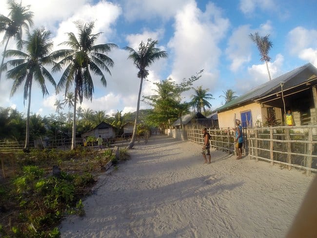 A sandy village path in Barangonan is lined with palm trees, wooden fences, and houses under a partly cloudy sky. Ideal for a break after an island hopping adventure, two people stand on the right side of the path.