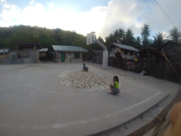 Two people sit on a concrete court surrounded by a pile of dried fish, reminiscent of a scene from Barangonan. In the background, houses and trees stand tall beside a weathered basketball hoop—much like stops on an island-hopping boat tour.