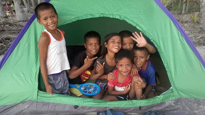 A group of smiling children, some inside and some standing outside a green and purple tent, enjoy their camping adventure. One child holds a toy, and another waves at the camera. The backdrop features trees, capturing the joyful spirit of outdoor exploration in a setting reminiscent of Palawan.