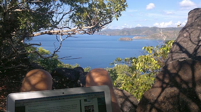 A person sits with a laptop in a scenic outdoor setting, surrounded by trees and rocks, overlooking the ocean. It's a perfect spot to plan an island hopping adventure or book a boat tour through the breathtaking landscapes of Palawan.