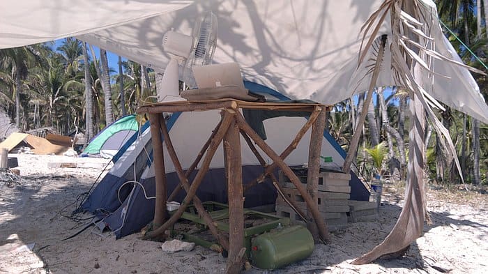 A makeshift outdoor office on a tropical Palawan beach features a tent, table, laptop, and fan under a tarp, perfectly blending work with the allure of island hopping adventures as boat tours await nearby.