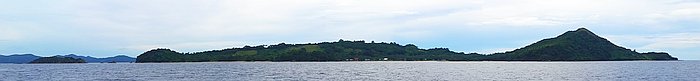 Panoramic view of a lush, green island with a prominent hill, ideal for island hopping and surrounded by calm ocean waters under a cloudy sky.