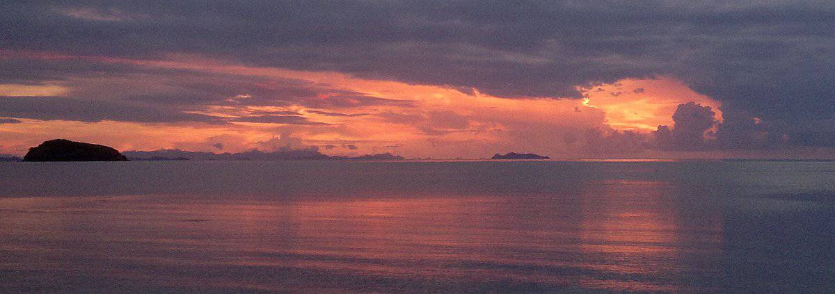Experience a serene sunset on a boat tour, as vibrant orange and pink hues reflect on the water, framed by dark clouds and distant silhouetted islands.