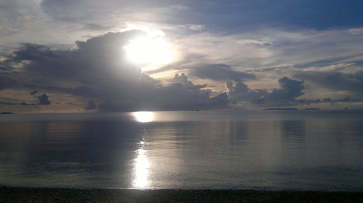 Sunrise light filters through clouds over a calm sea in Palawan, reflecting on the water with a distant horizon and subtle island silhouettes, as a boat tour quietly floats by.