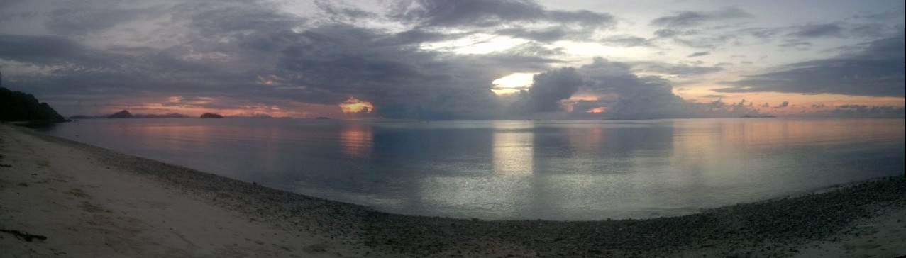A serene beach at sunrise in Palawan, with a cloudy sky reflecting on calm ocean waters and a rocky shoreline.